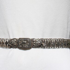 May include: A silver chain belt with a decorative, ornate buckle. The buckle is made of silver and has a heart-shaped design.