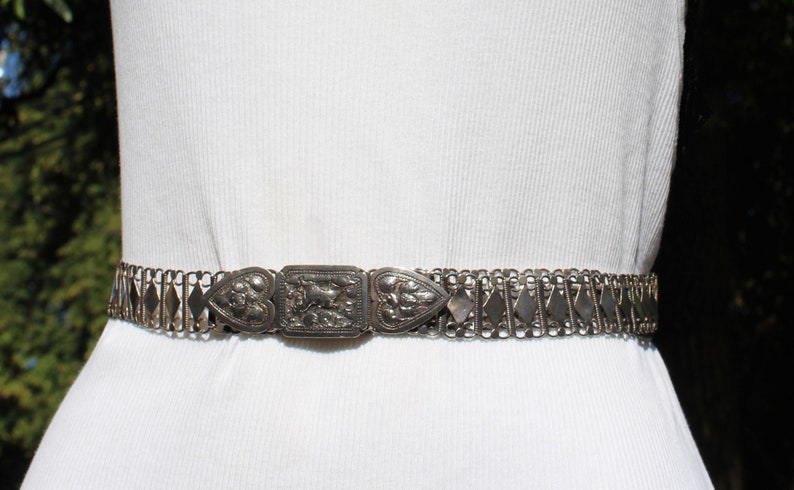 May include: A silver chain belt with a decorative, ornate buckle. The buckle is made of silver and has a heart-shaped design.
