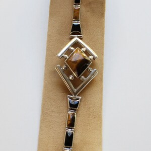 May include: A silver bracelet with a geometric design featuring a large tiger's eye gemstone. The bracelet is made up of alternating black and tiger's eye stones.