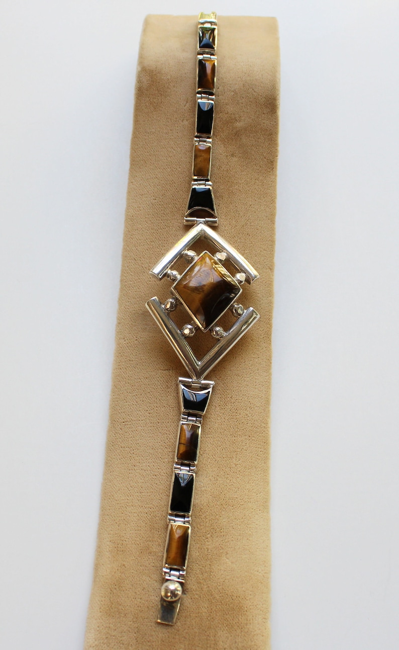 May include: A silver bracelet with a geometric design featuring a large tiger's eye gemstone. The bracelet is made up of alternating black and tiger's eye stones.