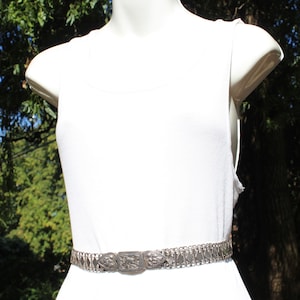 May include: A silver chain belt with an ornate, rectangular buckle. The belt is worn on a white tank top.