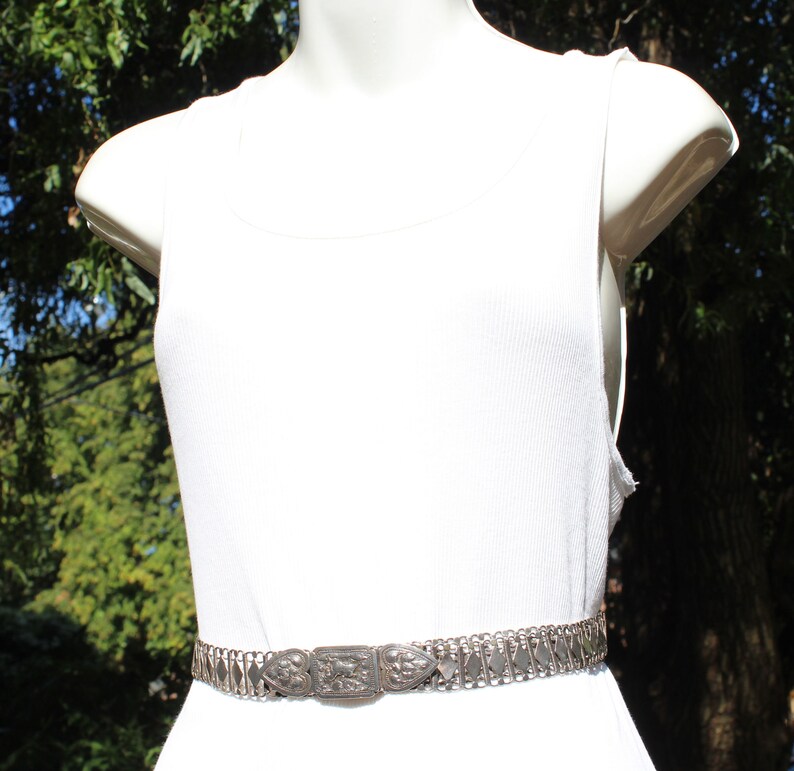 May include: A silver chain belt with an ornate, rectangular buckle. The belt is worn on a white tank top.