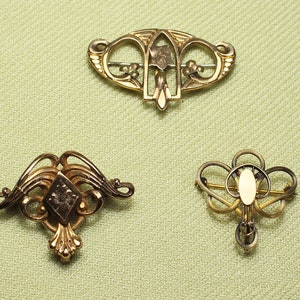 May include: Three gold-toned brooches with intricate designs. The brooches are on a light green fabric background.