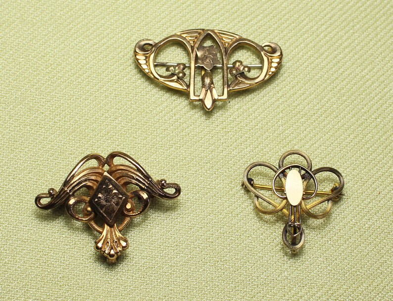 May include: Three gold-toned brooches with intricate designs. The brooches are on a light green fabric background.