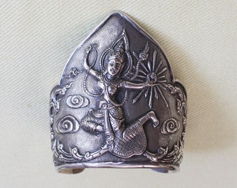 May include: Silver bracelet with an intricate design of a dancing figure with swirling clouds and a sunburst. The figure is wearing a traditional costume and has a crown on its head.