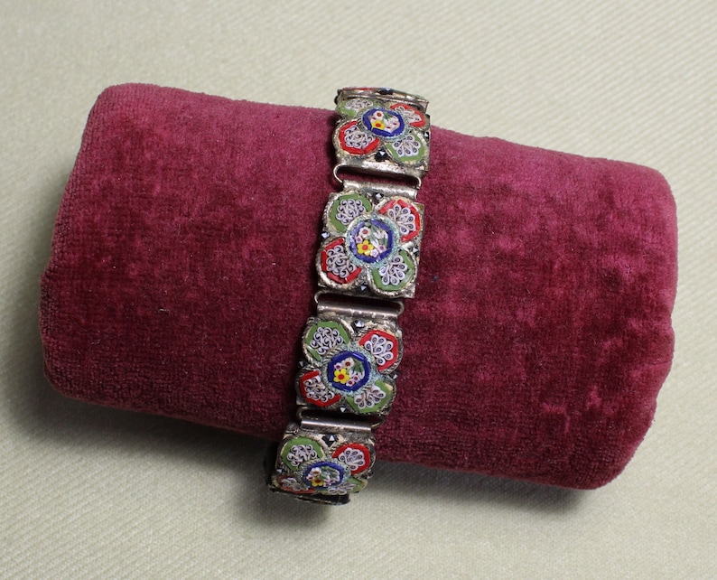 May include: A silver bracelet with a floral design made of colorful mosaic tiles. The bracelet is on a red velvet surface.