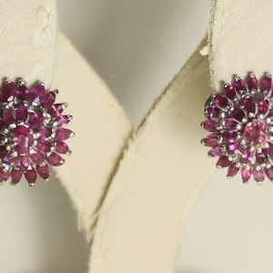 以下が含まれることがあります： 花型のスタッドピアス。各イヤリングは、中央の丸いピンク色の宝石の周りに配置された、小さな楕円形の濃いピンク色の宝石のクラスターを備えています。 イヤリングは銀色の金属にセットされています。