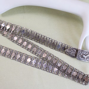 May include: A silver chain belt with a decorative clasp. The belt is made of interlocking links and has a repeating pattern of diamond-shaped pieces. The clasp is ornate and features a floral design.