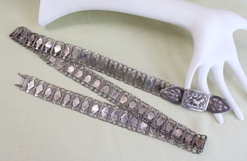 May include: A silver chain belt with a decorative clasp. The belt is made of interlocking links and has a repeating pattern of diamond-shaped pieces. The clasp is ornate and features a floral design.