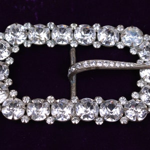 May include: A silver-toned belt buckle with a decorative design of clear rhinestones. The buckle is oval-shaped and has a curved metal clasp.