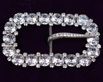 May include: A silver-toned belt buckle with a decorative design of clear rhinestones. The buckle is oval-shaped and has a curved metal clasp.