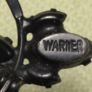 May include: Close-up of a black metal brooch with a floral design. The brooch has a silver oval tag with the word "WARNER" embossed on it.