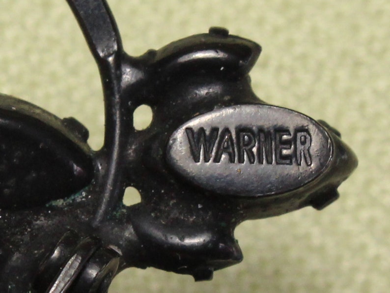 May include: Close-up of a black metal brooch with a floral design. The brooch has a silver oval tag with the word "WARNER" embossed on it.