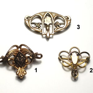 May include: Three gold-toned brooches with intricate designs. The top brooch features a white flower with a gold center. The left brooch has a diamond-shaped design with a gold center. The right brooch has a circular design with a white center.