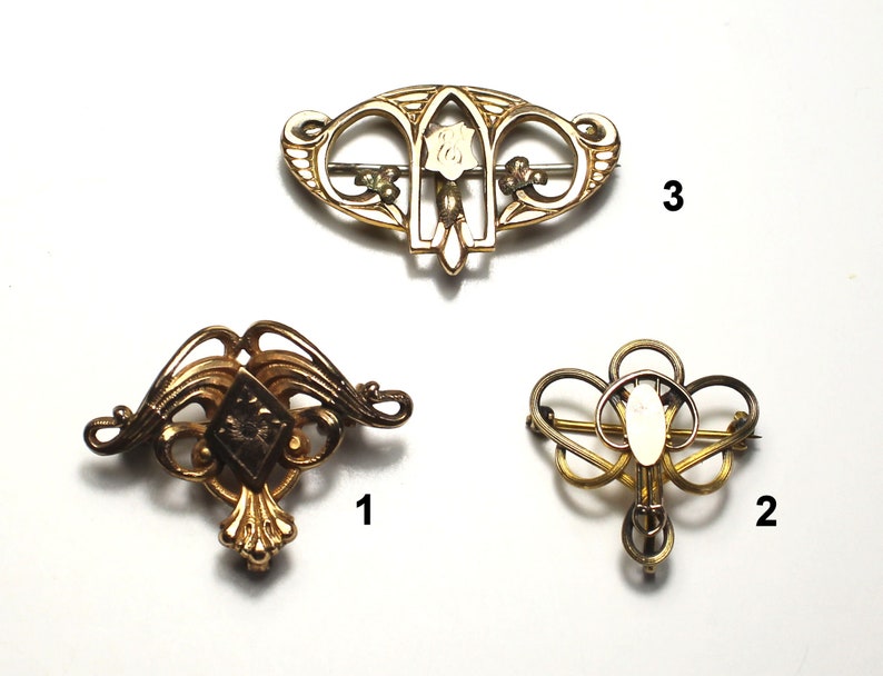 May include: Three gold-toned brooches with intricate designs. The top brooch features a white flower with a gold center. The left brooch has a diamond-shaped design with a gold center. The right brooch has a circular design with a white center.