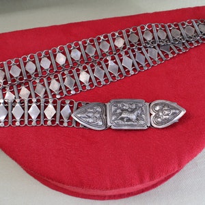 May include: A silver chain belt with a decorative clasp. The belt is made of interlocking diamond-shaped links and has a heart-shaped buckle with intricate engravings.