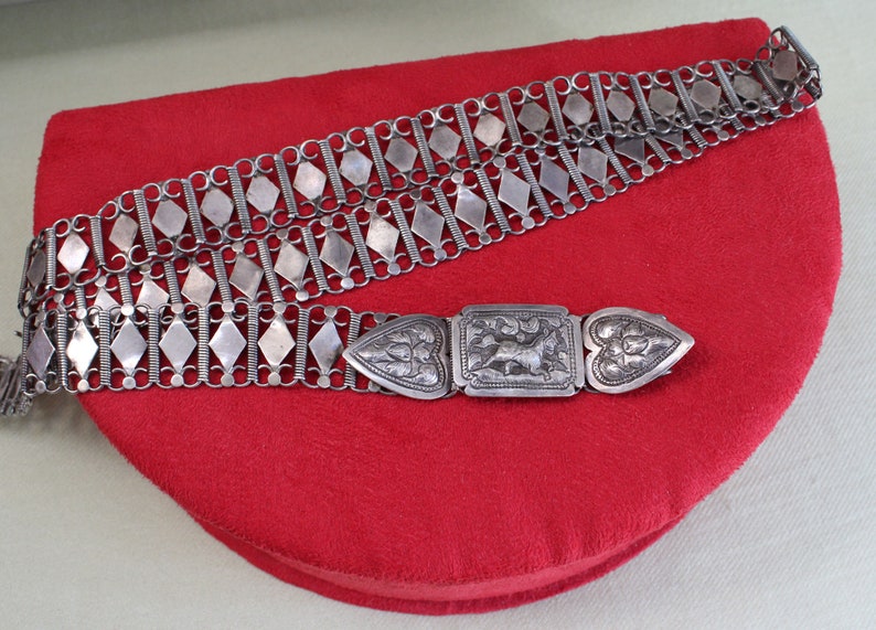 May include: A silver chain belt with a decorative clasp. The belt is made of interlocking diamond-shaped links and has a heart-shaped buckle with intricate engravings.