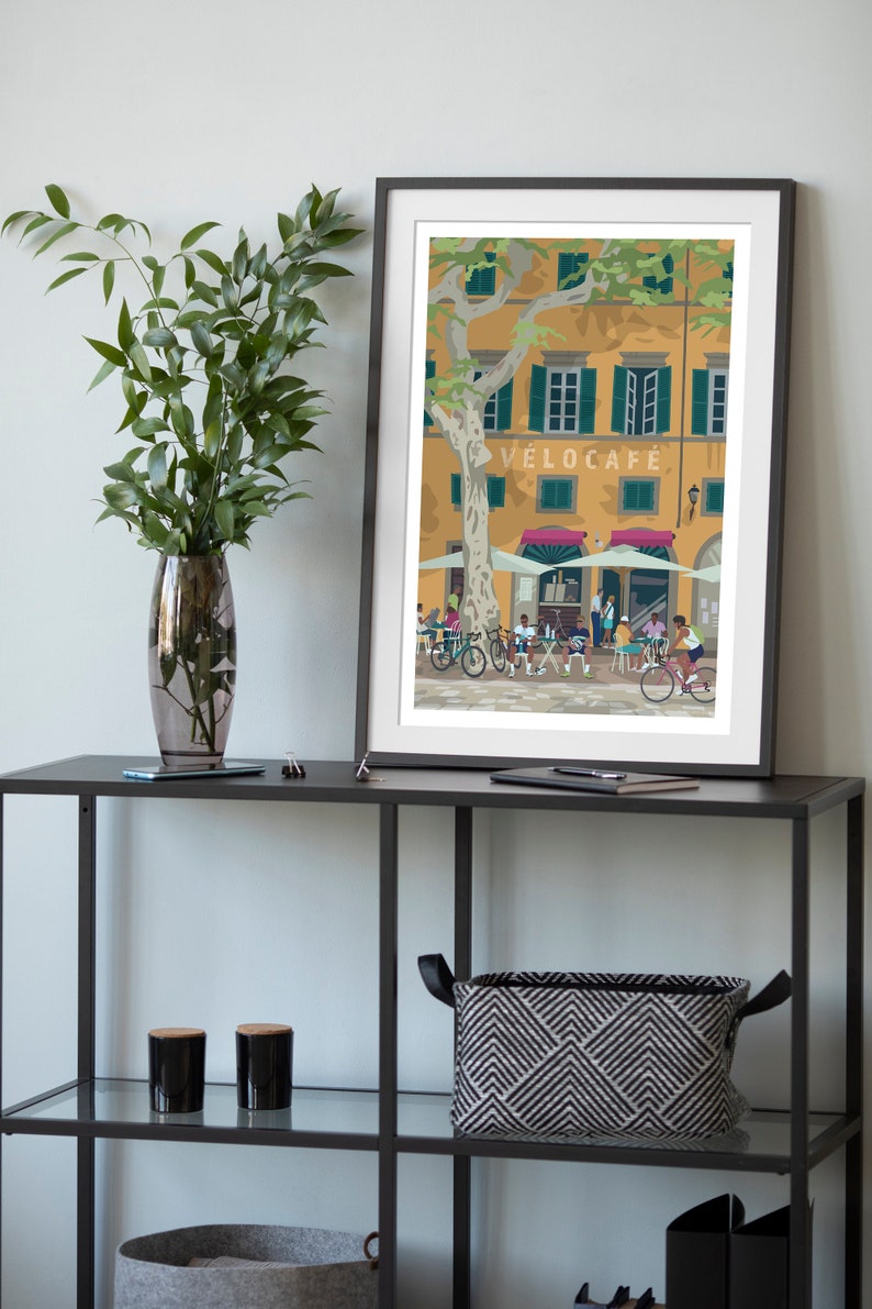 Velocafe cycling themed print showed framed and in a room environment on a black shelf with a vase and plant.