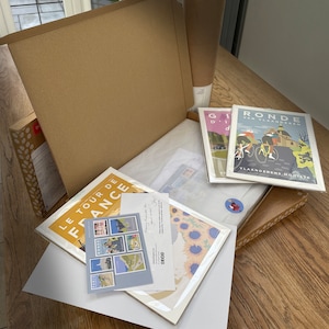 May include: Example packaging for cycling posters. The posters feature illustrations of cyclists and text in Dutch and French. The posters are rolled up and placed in a cardboard box with tissue paper.