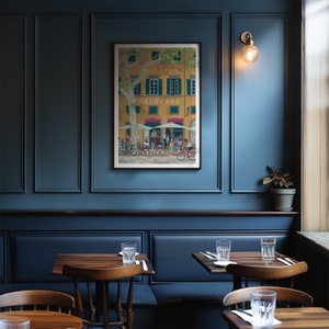 May include: A framed art print depicting a cafe scene with cyclists. The artwork features a yellow building with green shutters and the text "VELOCAFE". Cyclists are shown in front of the cafe, with a large tree providing shade. The print is displayed on a dark blue wall.