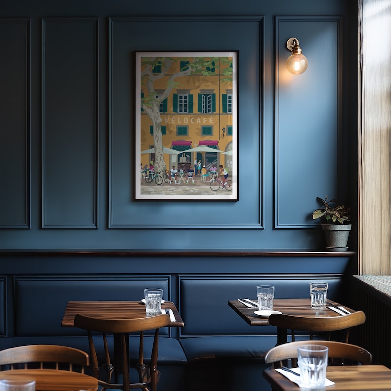 May include: A framed art print depicting a cafe scene with cyclists. The artwork features a yellow building with green shutters and the text "VELOCAFE". Cyclists are shown in front of the cafe, with a large tree providing shade. The print is displayed on a dark blue wall.