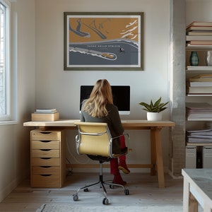 May include: A framed poster of Passo dello Stelvio, a mountain pass, hangs above a wooden desk. A person sits at the desk in front of a computer. A wooden file cabinet with five drawers is to the left of the desk. A potted plant sits on the desk.