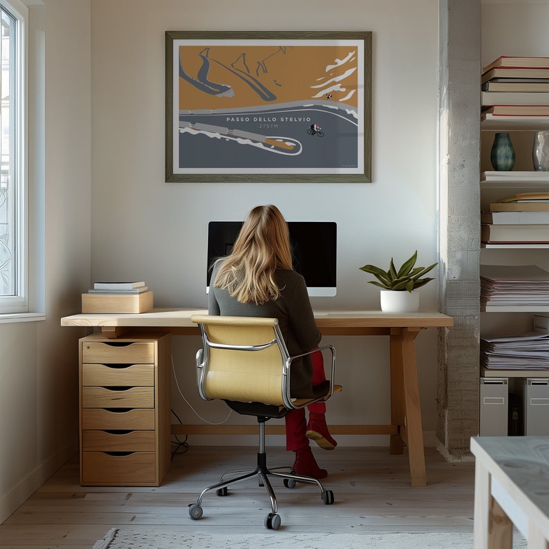May include: A framed poster of Passo dello Stelvio, a mountain pass, hangs above a wooden desk. A person sits at the desk in front of a computer. A wooden file cabinet with five drawers is to the left of the desk. A potted plant sits on the desk.