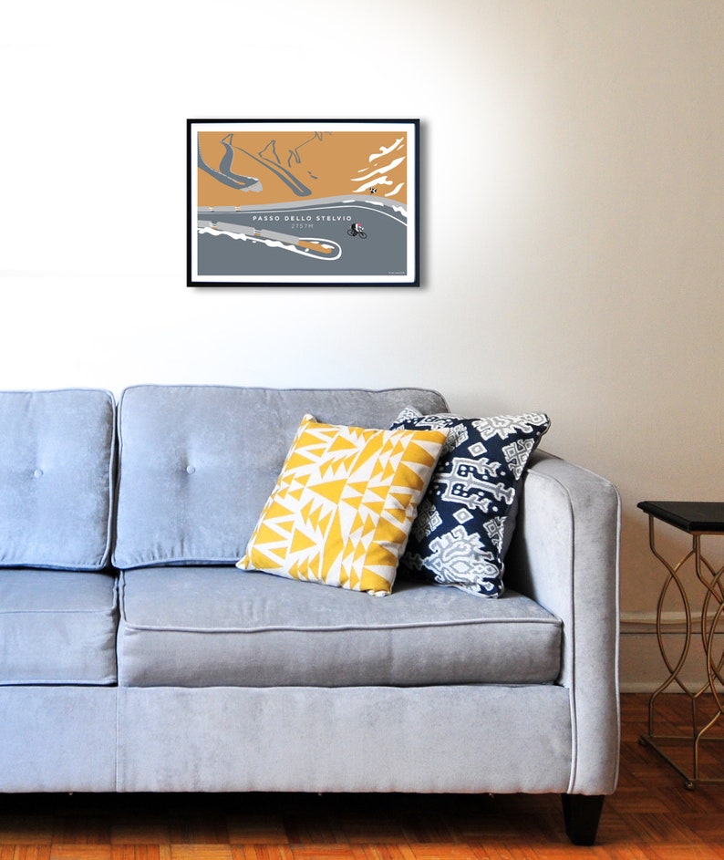 Room setting with grey sofa and wall art Passo Dello Stelvio  - Almost at the Summit Cycling Print. Simplistic style mainly grey and mustard colour design, looking down on cyclist ascending the hairpin bends on Stelvio Pass in Italy.