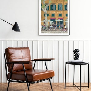 Vélo Café cycling themed print shown framed on the wall with a leather chair and wooden floor, black side table and fluffy rug.