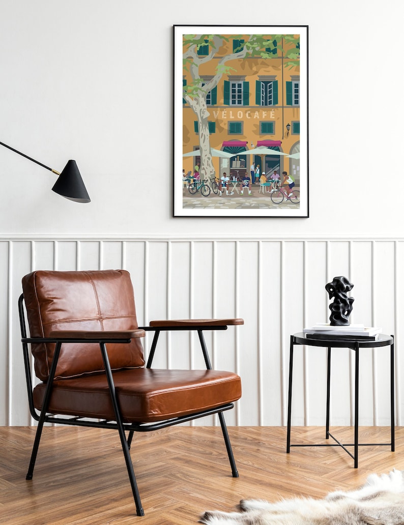 Vélo Café cycling themed print shown framed on the wall with a leather chair and wooden floor, black side table and fluffy rug.