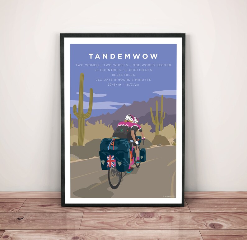 Framed art print leaning against a wall. TandemWoW Guinness World Record Holders Cycling Print showing the women tandem cyclists riding through a desert with cacti and mountains on their world record breaking challenge.