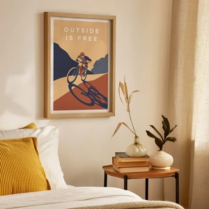 May include: A framed poster with a minimalist design featuring a person riding a bicycle on a mountain path. The poster has the text "OUTSIDE IS FREE" in white letters on a light brown background.