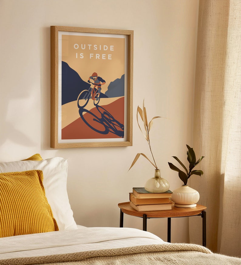 May include: A framed poster with a minimalist design featuring a person riding a bicycle on a mountain path. The poster has the text "OUTSIDE IS FREE" in white letters on a light brown background.