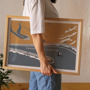 May include: A framed poster with a cycling illustration of Passo dello Stelvio. The artwork has a minimalist style with a cyclist on a winding road, set against a mustard yellow and grey background. The frame is made of light-coloured wood.