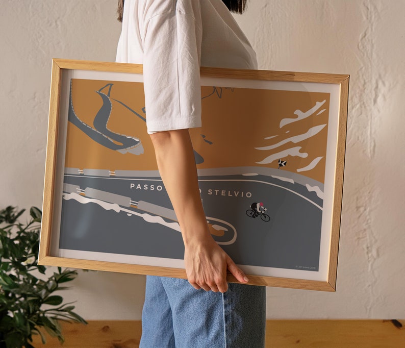 May include: A framed poster with a cycling illustration of Passo dello Stelvio. The artwork has a minimalist style with a cyclist on a winding road, set against a mustard yellow and grey background. The frame is made of light-coloured wood.