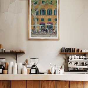 May include: A framed art print featuring a cafe scene with cyclists. The artwork depicts a yellow building with green shutters, a tree, and people at tables. The cafe is labeled "VELOCAFE". The print is in a gold frame.