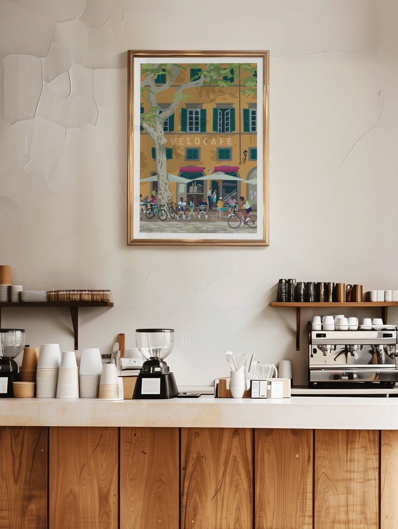 May include: A framed art print featuring a cafe scene with cyclists. The artwork depicts a yellow building with green shutters, a tree, and people at tables. The cafe is labeled "VELOCAFE". The print is in a gold frame.