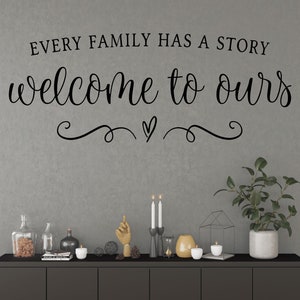 May include: A black and white wall decal with the text "Every family has a story, welcome to ours" with a heart and swirls underneath. The decal is on a gray wall above a black shelf with candles, a gold hand sculpture, and a potted plant.