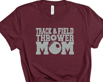 May include: Burgundy t-shirt with silver glitter text that reads "Track & Field Thrower Mom" with a silver glitter shot put graphic.