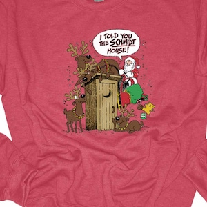 May include: Red sweatshirt with a humorous Christmas graphic. The graphic features Santa Claus, reindeer, and a green Grinch-like character gathered around an outhouse. Text in a speech bubble reads, "I told you the Schmidt House!"