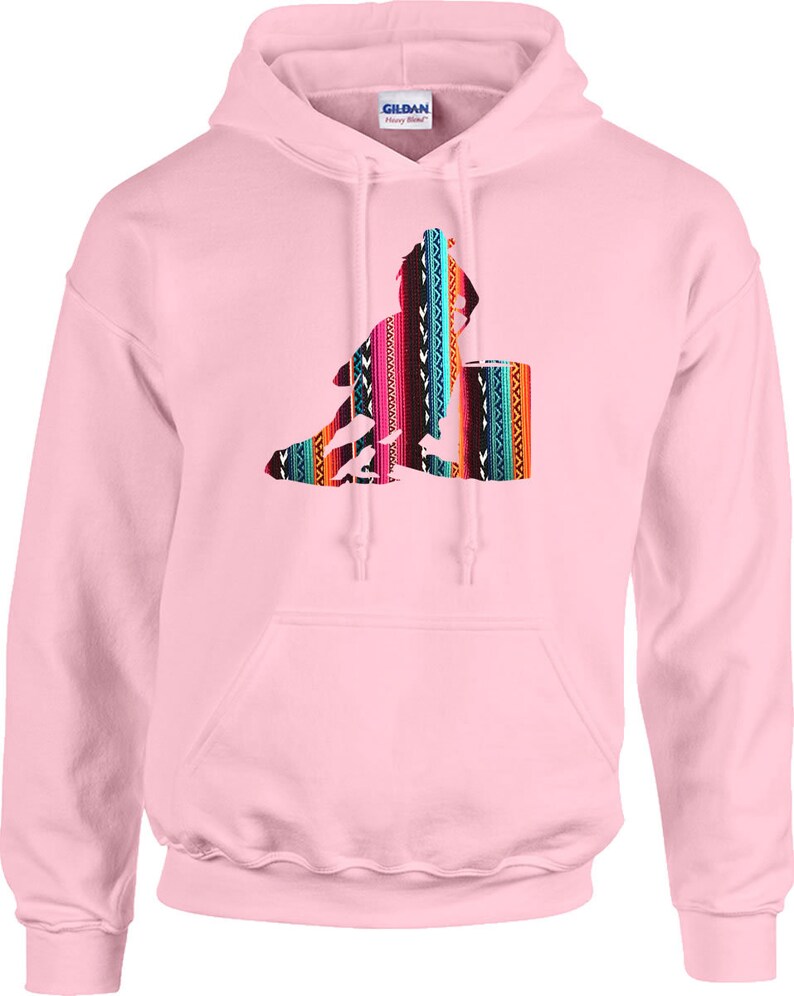 May include: Pink hooded sweatshirt featuring a colourful, geometric design of a person sitting on a barrel. The design is reminiscent of a traditional Mexican blanket.