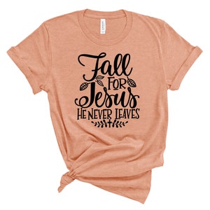 May include: A peach-colored t-shirt with a black graphic that reads "Fall for Jesus He Never Leaves". The graphic includes stylized leaves.