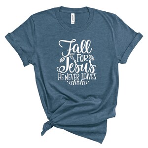 May include: A blue t-shirt with white text that reads "Fall for Jesus He Never Leaves". The text is in a cursive font and is surrounded by small leaf designs.