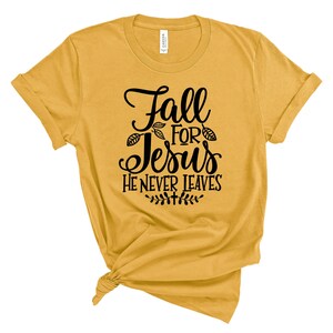 May include: A mustard yellow t-shirt with the text "Fall for Jesus He Never Leaves" in black lettering. The text is surrounded by small leaf designs.