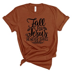 May include: A brown t-shirt with the text "Fall for Jesus He Never Leaves" in black lettering. The text is surrounded by small leaf designs.