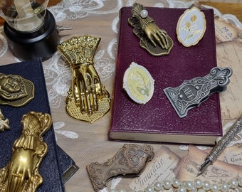 Może przedstawiać: Kolekcja ozdobnych metalowych podpór do książek i spinaczy do papieru w odcieniach złota i srebra. Podpory do książek mają misterne wzory dłoni i kwiatów. Spinacze do papieru mają ozdobne wzory i zawierają napis "Art".