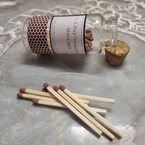 May include: A small glass bottle filled with matches, with a cork stopper. The label reads "L.A. Ilay Treasure Matches". Several matches are scattered in front of the bottle. The match heads are a reddish-brown color.