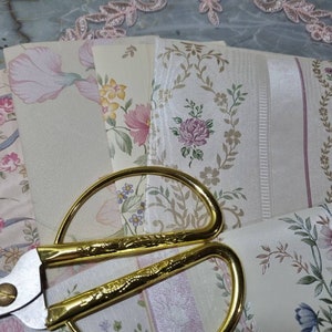 May include: A pair of gold-coloured scissors with a silver blade are lying on top of several pieces of floral patterned wallpaper. The wallpaper has a light background with pink, green, and brown floral designs.