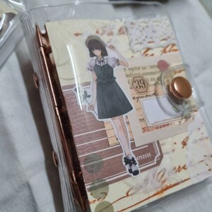 May include: A clear plastic binder with a brown and white patterned paper insert. The insert features a drawing of a girl in a black dress and a hat, along with a number "39" and other decorative elements.