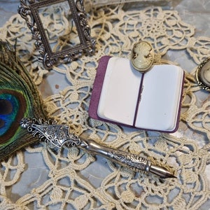 Può includere: Una penna stilografica in piuma di pavone argentata e bianca con un manico decorativo, un piccolo taccuino viola e due spille cameo su un pizzo all'uncinetto bianco.